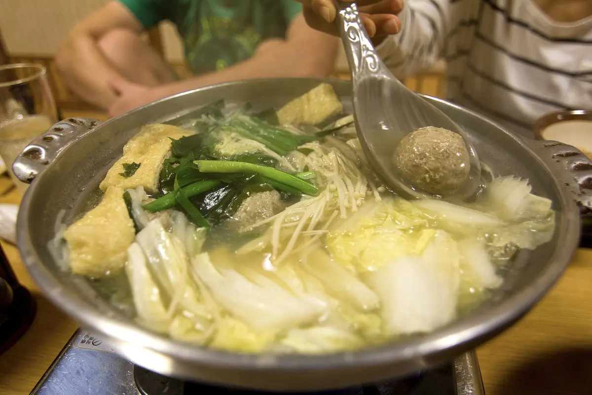 A steaming silver pot of chankonabe with tofu pouches, leafy greens, enoki mushrooms and a meatball on a ladle