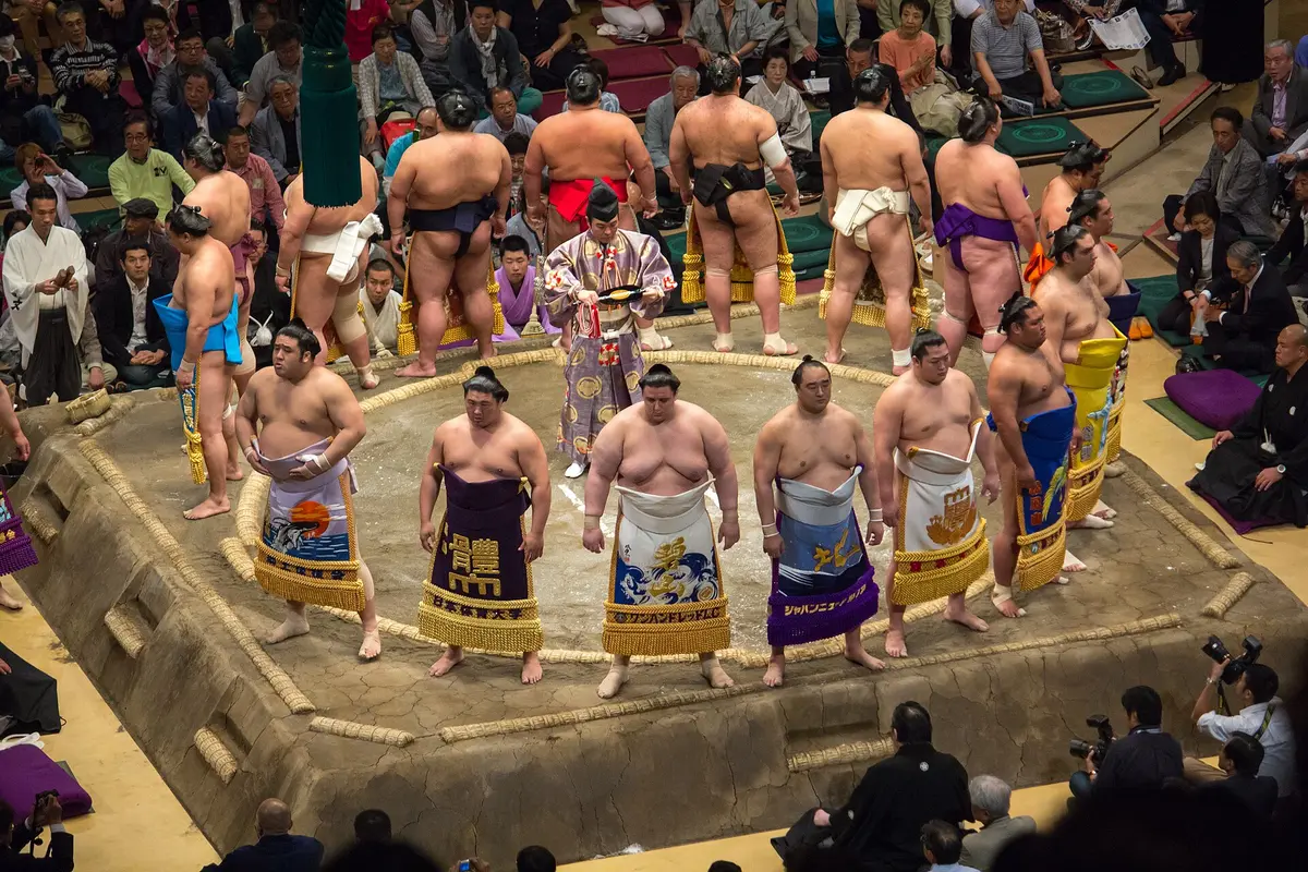 Wrestlers in decorated ceremonial aprons standing in a ring on a raised clay platform marked with straw bales, during a ring-entering ceremony